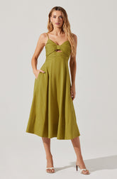 A woman is wearing a sleeveless, olive-green dress with spaghetti straps. The dress has a fitted bodice with a large, knotted detail at the center of the bust, creating a keyhole cutout below the knot. The skirt is full and flares out to mid-calf length, with a wide hem at the bottom. She completes the look with beige, open-toe high-heeled sandals.